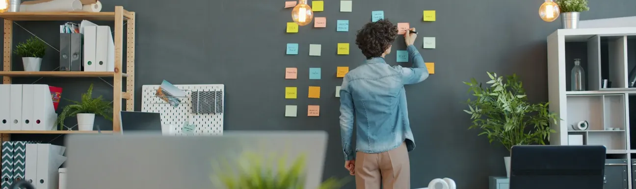 Personne debout devant un mur, écrivant sur des notes autocollantes disposées en grille dans un espace de travail.