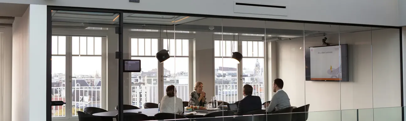 Salle de réunion vitrée avec une table centrale et plusieurs personnes assises, dans un espace de travail professionnel lumineux.