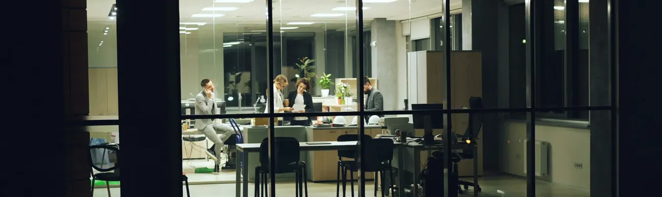 Office interior at night with people working, viewed through large windows.