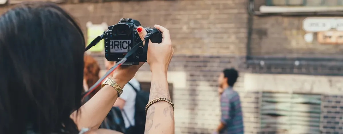 A photographer uses a digital SLR camera to capture an urban scene. People are seen moving in the background, and there is a brick environment.