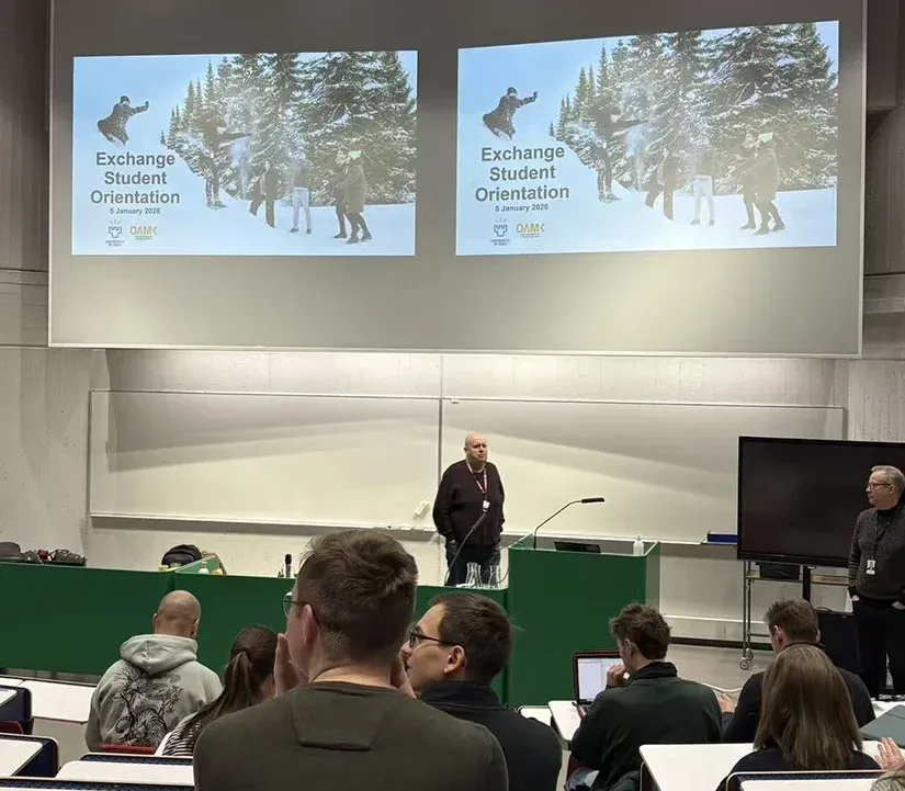 Un groupe d'étudiants assiste à une séance d'orientation pour les étudiants d'échange. À l'arrière-plan, deux écrans affichent un diaporama avec des images d'activités hivernales, telles que des personnes dans la neige. L'environnement semble académique, avec un présentateur debout devant la classe. Les étudiants prennent des notes sur des ordinateurs portables ou des documents.