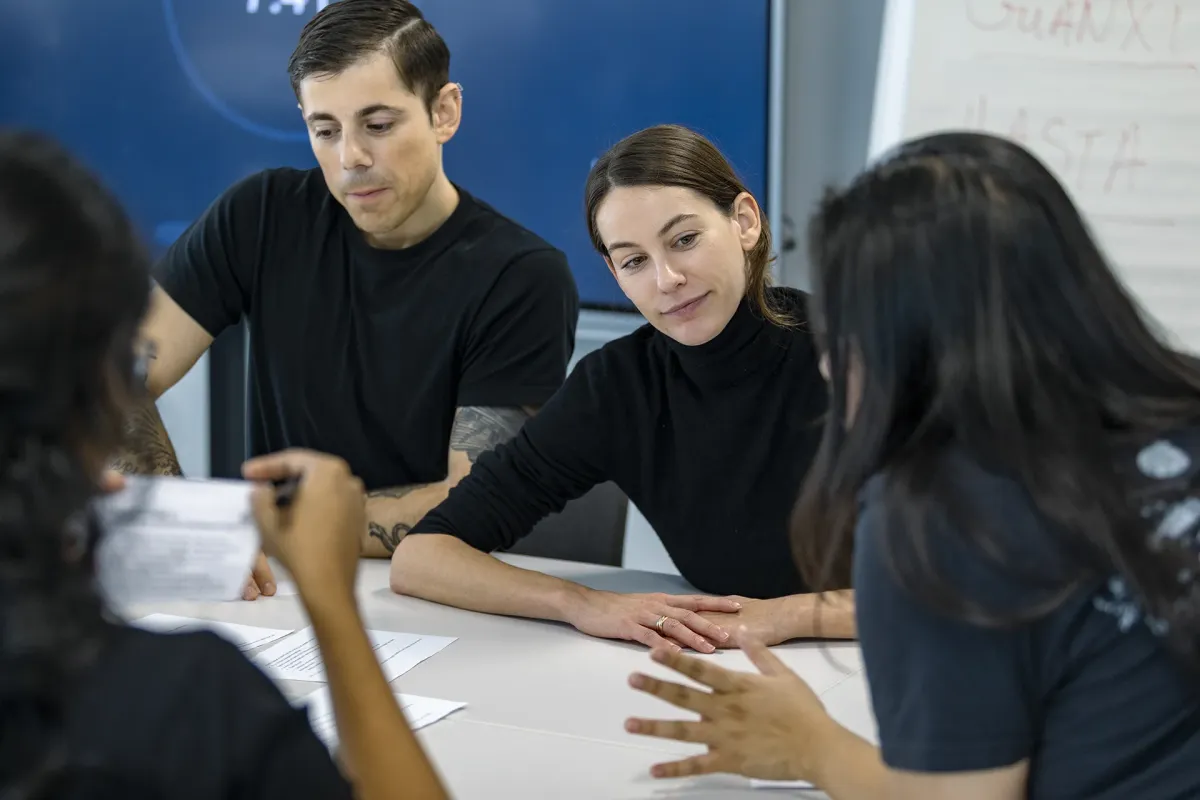 Réunion de groupe dans un bureau, avec plusieurs personnes discutant autour d'une table, des documents et un tableau en arrière-plan.