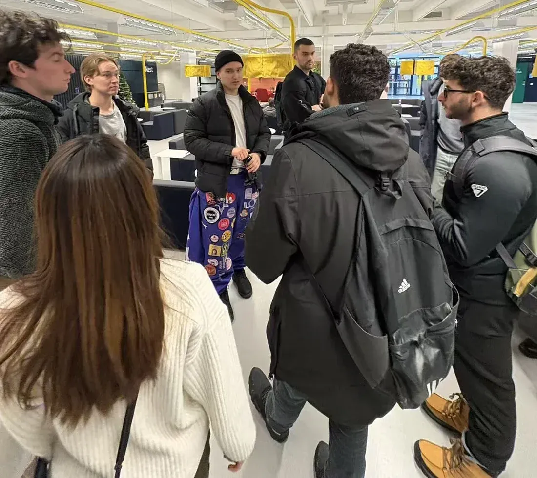 Un groupe de personnes est rassemblé dans un espace intérieur moderne, probablement dans un établissement éducatif. Ils semblent engagés dans une conversation animée, certains ayant des vêtements décontractés et une atmosphère conviviale. L'espace est éclairé de manière brillante, avec des meubles contemporains visibles en arrière-plan.