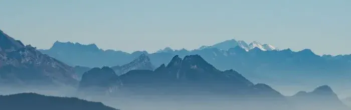 Chaîne de montagnes bleutées dans la brume au lever du soleil, avec sommets alpins visibles à l’horizon.