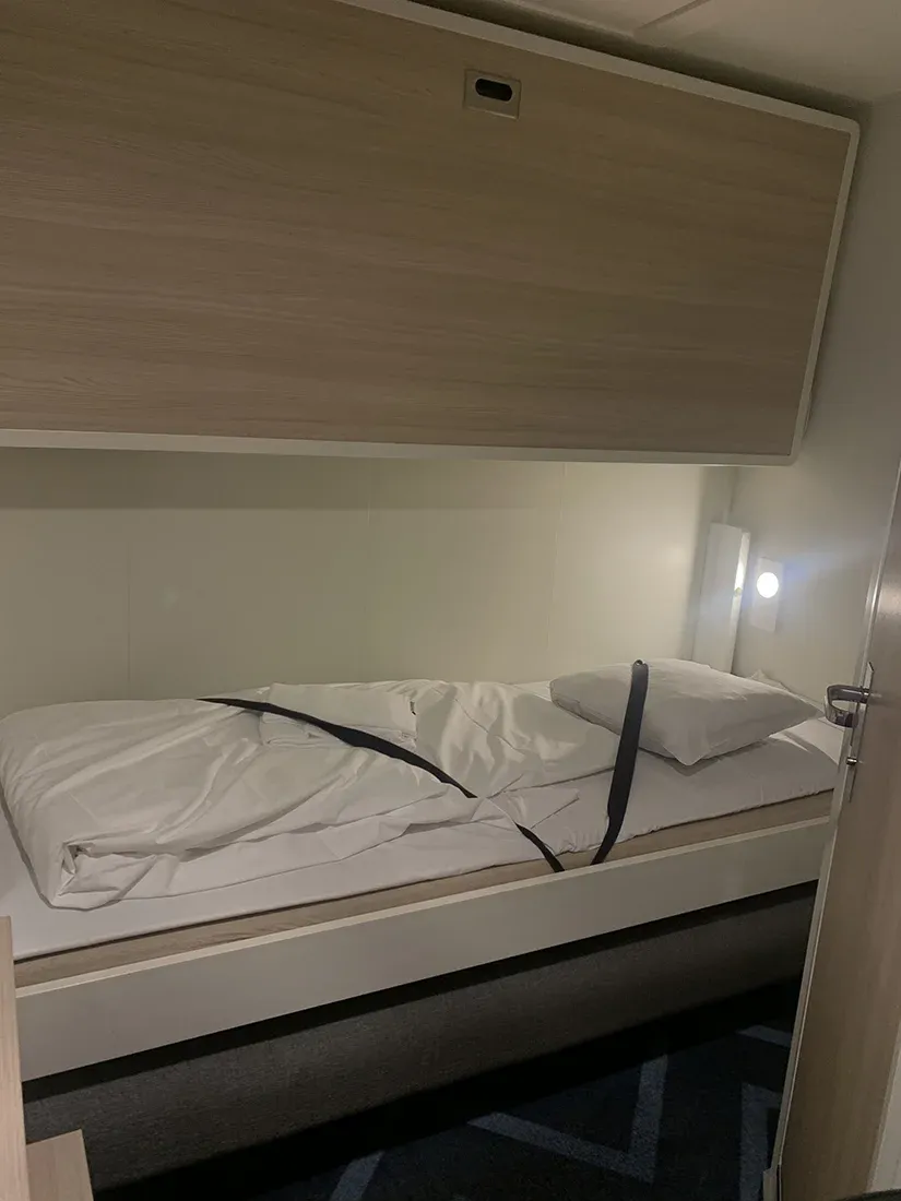 Une petite cabine avec un lit simple fait, un drap retenu par des sangles et un éclairage mural, montrant un décor minimaliste.