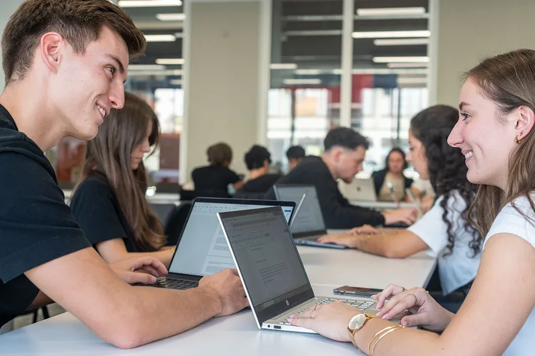Étudiants d'emlyon travaillant ensemble sur leurs ordinateurs portables dans un espace de travail collaboratif du campus, concentrés sur un projet académique.