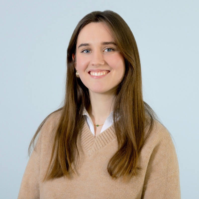 Portrait-style image of Manon Borie (emlyon student) wearing a beige sweater over a white shirt, standing against a plain light-colored background.