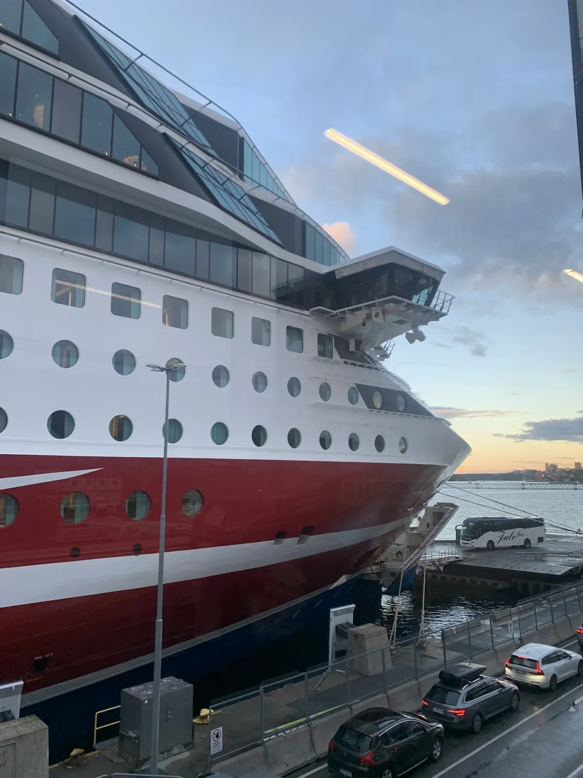 Large red and white ferry docked at the harbor, viewed from the terminal with cars parked nearby and the sea in the background at sunset.