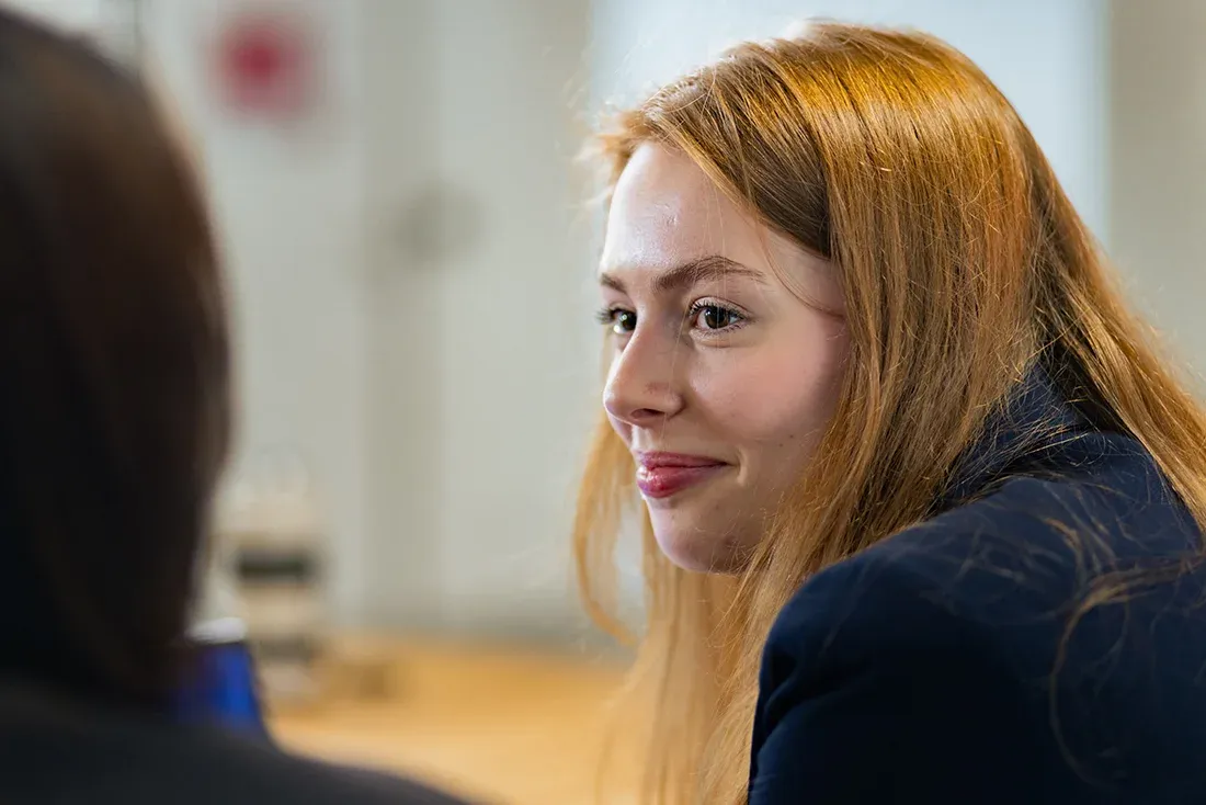 Person seated in a professional environment, viewed from the side, engaged in a conversation or interview with another person
