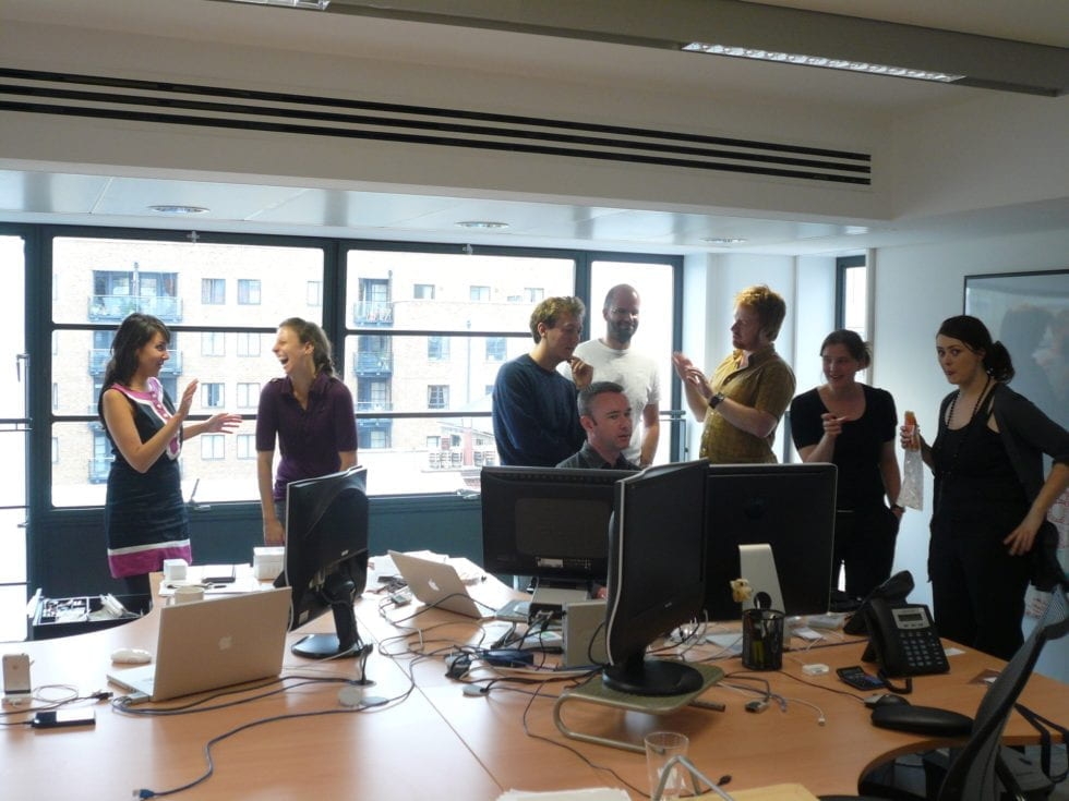 Groupe de personnes réunies dans un bureau autour de postes de travail et d’ordinateurs, lors d’une discussion informelle.