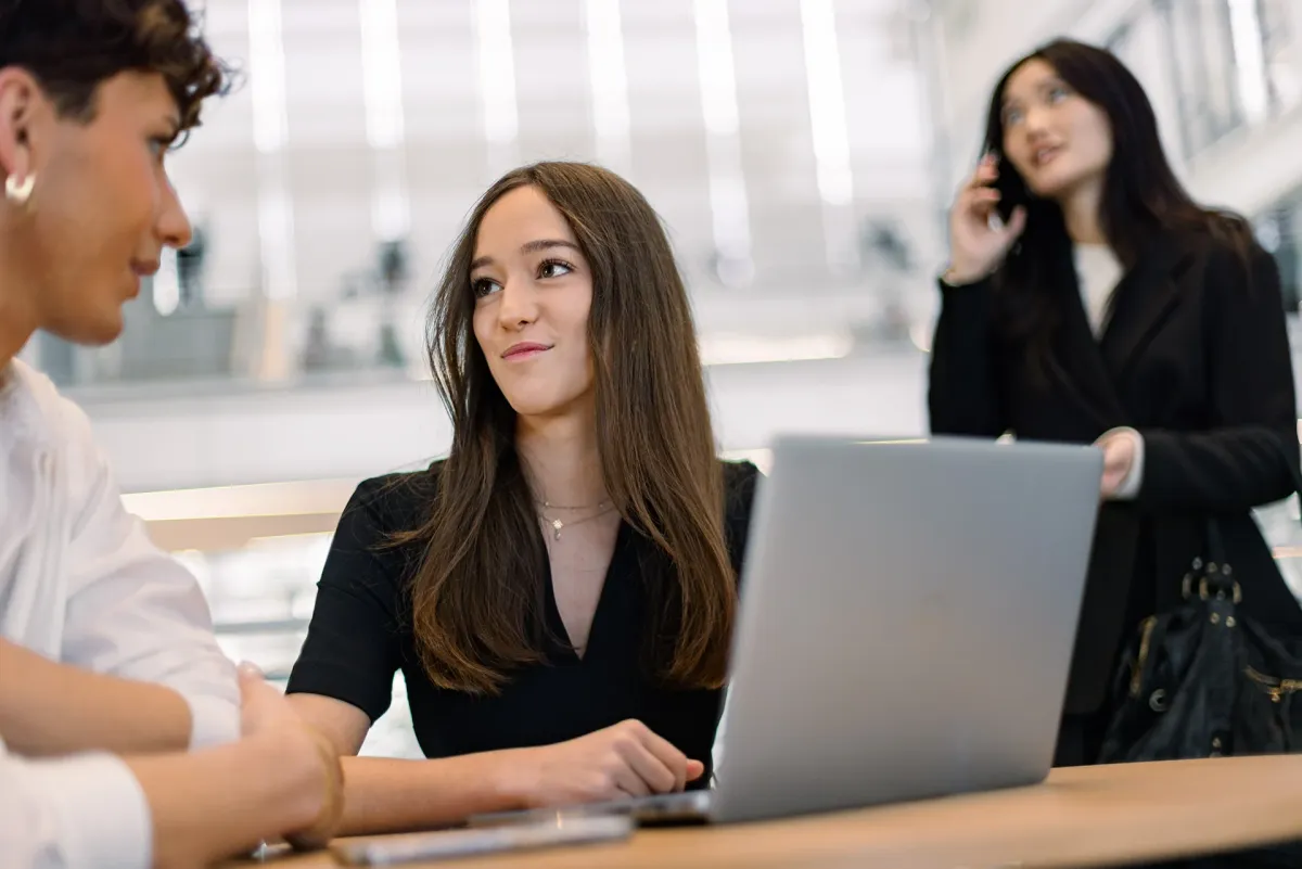  Trois personnes travaillent ensemble dans un espace moderne de formation. Une personne utilise un ordinateur portable sur une table, tandis que deux autres échangent à proximité, dans un environnement lumineux évoquant l’enseignement supérieur et l’alternance.