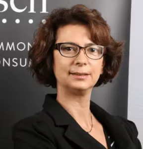 Portrait photographique d’une personne cadrée en buste, portant une veste sombre et des lunettes, devant un fond avec du texte partiellement visible.