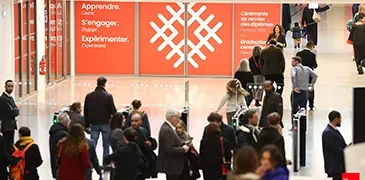 Personnes circulant dans le hall d’un campus universitaire, avec des panneaux institutionnels orange en arrière‑plan.
