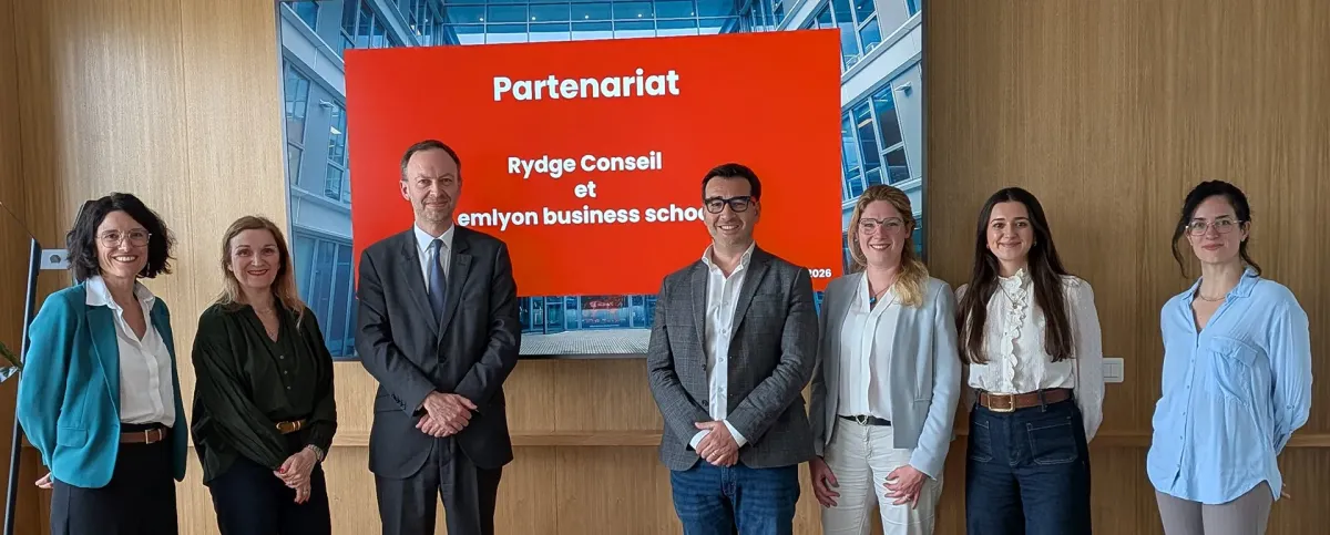 Un groupe de personnes se tient debout dans un espace intérieur, devant un écran affichant le texte "Partenariat Rydge Conseil et emlyon business school "
