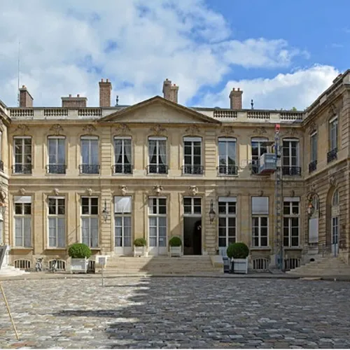 Cour intérieure d’un bâtiment historique avec façade en pierre, fenêtres symétriques et sol pavé sous un ciel partiellement nuageux.