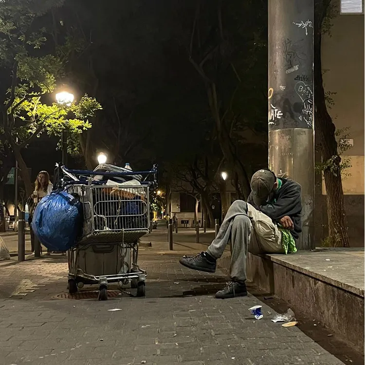 Personne assise sur un trottoir la nuit à côté d’un chariot rempli d’affaires, sous un lampadaire dans une rue urbaine.