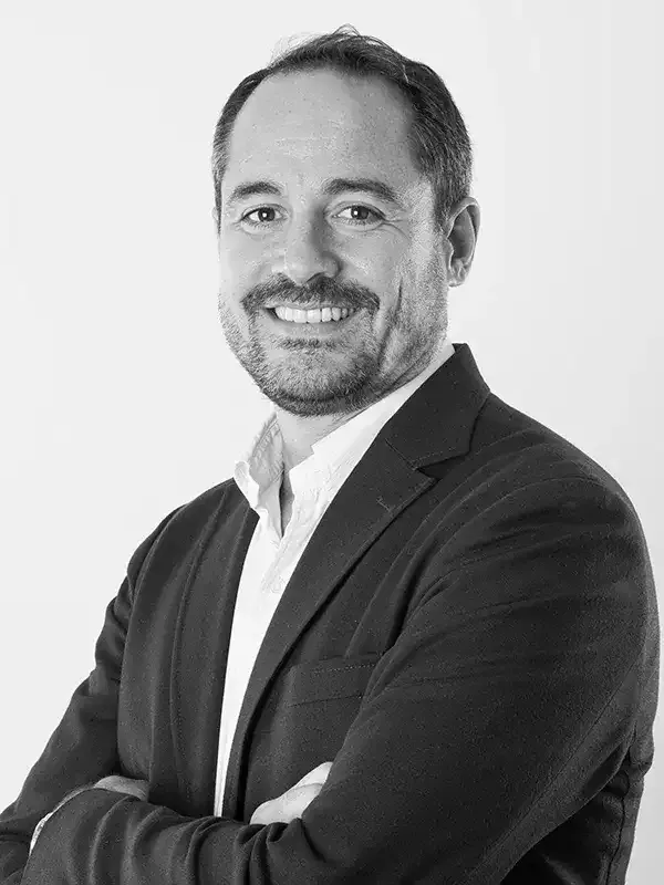 Portrait photographique en noir et blanc d’une personne cadrée en buste, portant une veste sombre sur une chemise claire, les bras croisés, sur un fond uni clair.