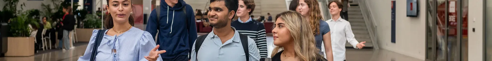 Un groupe d’étudiants et d’étudiantes d’emlyon marche dans le vaste atrium lumineux de l’école. L’espace présente de grandes baies vitrées, des passerelles, des escaliers et des éléments architecturaux rouges qui structurent le bâtiment. Le groupe avance au premier plan en discutant, tandis que d’autres personnes se déplacent à l’arrière-plan dans le cœur battant du campus.