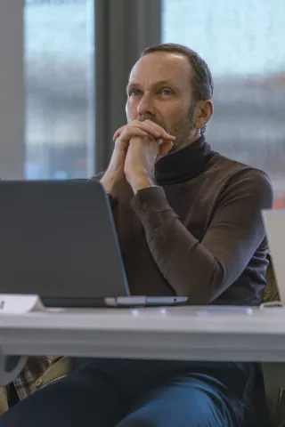 Un homme assis à une table de réunion, portant un pull col roulé brun, les mains posées sur son visage en signe de réflexion. Un ordinateur portable est visible devant lui, avec des éléments de bureau en arrière-plan