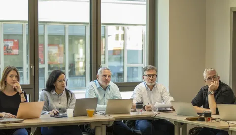 Réunion d'équipe dans une salle lumineuse, avec plusieurs participants assis autour d'une table, chacun devant un ordinateur portable. Des boissons sont également visibles sur la table.