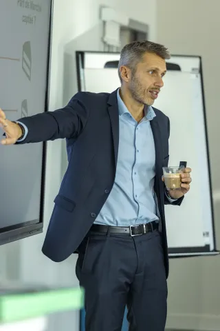 Un homme est en train de faire une présentation, vêtu d'un costume sombre et d'une chemise bleue. Il pointe vers un écran avec une main, tout en tenant une boisson dans l'autre. L'environnement semble officiel, probablement lors d'une réunion ou d'une conférence. La scène reflète une interaction professionnelle axée sur la communication d'idées ou d'informations.