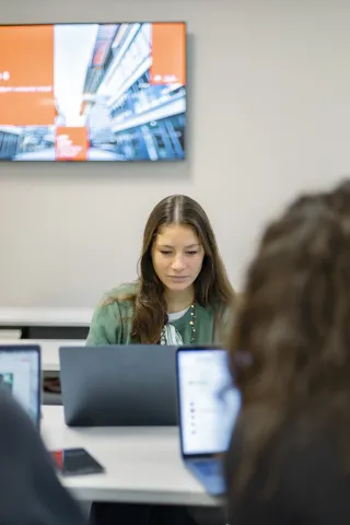 Réunion en cours avec une participante, portant un pull vert, devant des ordinateurs portables, avec un écran affichant une présentation en arrière-plan.