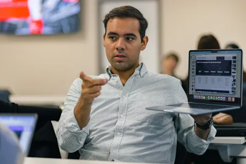 Une personne présentant un ordinateur portable tout en tenant une discussion, avec un graphique ou des informations visibles à l'écran. D'autres participants sont présents en arrière-plan, attentifs à la présentation.