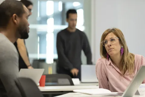Un groupe de personnes est engagé dans une conversation. Une femme, tournée vers un homme, semble partager une idée ou une question. D'autres participants se trouvent en arrière-plan, ajoutant à l'ambiance collaborative de la réunion.