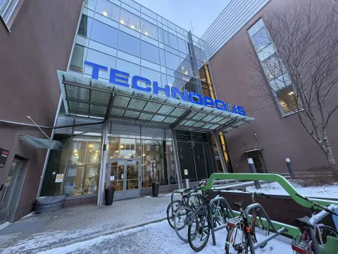 Le bâtiment devant vous est nommé "Technopolis". Il présente une architecture moderne avec une façade en verre réfléchissant et un toit en porte-à-faux. À l'entrée, on peut voir plusieurs vélos stationnés, suggérant que cet espace pourrait être fréquenté par des étudiants ou des professionnels. Le sol est légèrement enneigé, indiquant un climat froid.