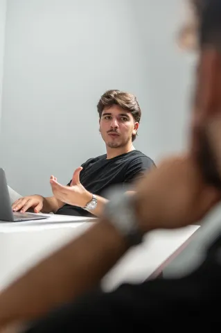 Une personne assise à une table, portant un t-shirt noir, en train de gesticuler et de parler. Sur la table, il y a un ordinateur portable. En arrière-plan, on aperçoit une autre personne dont la tête est légèrement floue. Les murs sont peints en gris clair.