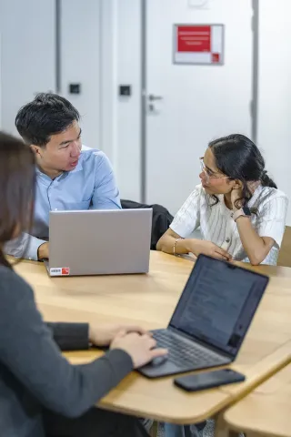 The image shows a group of people engaged in a meeting or discussion around a wooden table. Several laptops are open in front of them, indicating that they are working on tasks or sharing information. The workspace appears modern and well-lit, with a clean background. One person seems to be listening attentively while the others are conversing. Overall, the atmosphere suggests a collaborative environment.