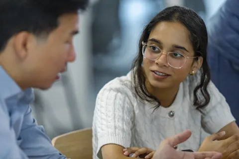 An exchange between two people around a table. One participant is turned toward the other, engaged in a discussion. The person on the right is wearing a textured white sweater with buttons and seems to be explaining a point. The environment is that of a modern office, conducive to collaboration.