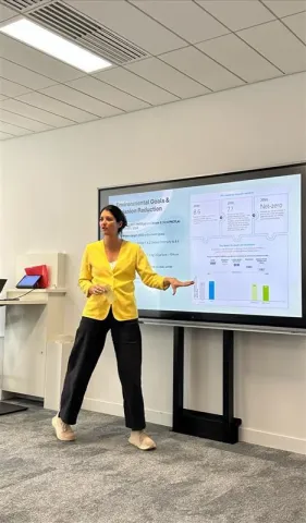 Erica, étudiante IMBA d'emlyon, donnant une présentation professionnelle dans une salle de réunion, devant un écran affichant des objectifs environnementaux et des graphiques.