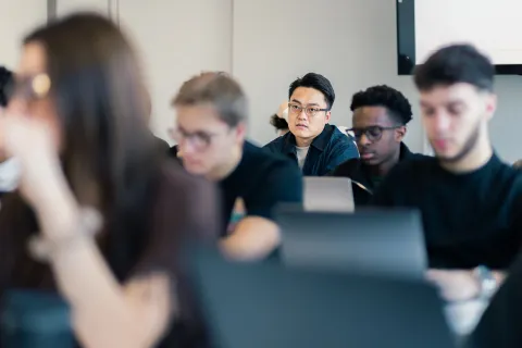 tudents sitting in a classroom during a lecture, focusing on laptops and listening to the instructor in a modern learning environment.