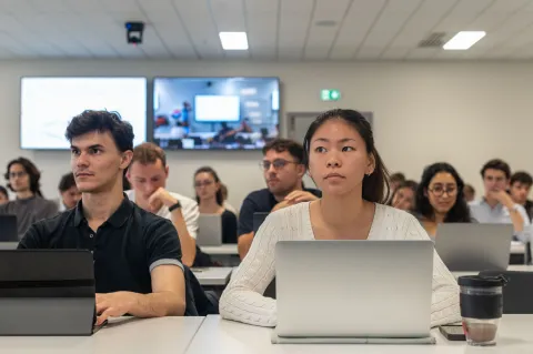 Students of emlyon attending a class in a modern classroom, working on laptops with digital screens displayed at the front.