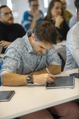 Participant de l’executive education d’emlyon prenant des notes pendant une session de formation, avec tablette numérique et ordinateur portable dans une salle de cours moderne.