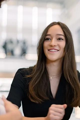Une femme portant un haut noir est assise à une table, regardant devant elle avec une expression concentrée. Elle a les cheveux longs et bruns, et porte des colliers délicats. En arrière-plan, un environnement de bureau moderne est visible avec des lumières et des éléments architecturaux lumineux.