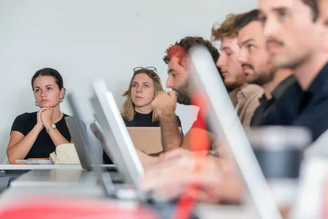Un groupe de personnes assises à une table dans une salle de réunion. Certaines personnes portent des vêtements noirs, et il y a plusieurs ordinateurs portables ouverts devant elles. L'ambiance semble être celle d'une discussion ou d'une présentation.