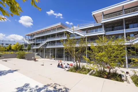 Vue du campus emlyon business school à Lyon, montrant un bâtiment moderne à plusieurs niveaux avec terrasses et passerelles extérieures, entouré d’arbres et d’espaces paysagers. Au premier plan, quelques personnes sont assises sur une esplanade en plein soleil.