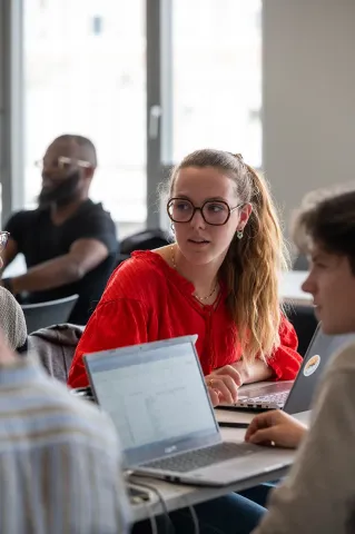 Student in a classroom working on a laptop during an in‑person lesson, in a bright room with large windows.
