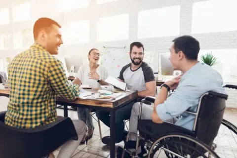 A group of people gathered around a work table in a bright space, discussing and sharing documents, including one person using a wheelchair.