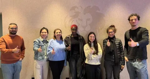 Un groupe d'étudiants et étudiants se tient devant un mur décoré avec le logo du lion d'emlyon. Tous montrent un pouce levé, exprimant une attitude positive. Les membres du groupe portent divers vêtements, allant de décontracté à plus habillé.