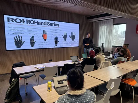 Une présentation en cours dans une salle de classe moderne, avec un intervenant devant un écran affichant des informations sur des gants robotiques. Des étudiants attentifs sont assis à des tables, chacun ayant un ordinateur portable devant eux. L'atmosphère est dynamique, favorisant l'échange d'idées et l'apprentissage.