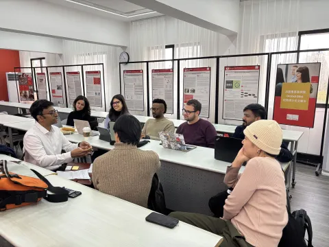 Un groupe d'étudiants en discussion active dans une salle de classe, entourés d'affiches académiques. Ils échangent des idées tandis que certains prennent des notes et d'autres consultent leurs ordinateurs portables. L'ambiance est collaborative et interactive, favorisant un apprentissage dynamique.