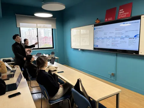 Un enseignant présente des informations sur un tableau interactif dans une salle de classe d'une école de commerce. Des étudiants assis à des tables utilisent des ordinateurs portables, tandis qu'une fenêtre laisse entrer la lumière naturelle. Les murs sont peints en bleu et des affiches de l'école sont accrochées.
