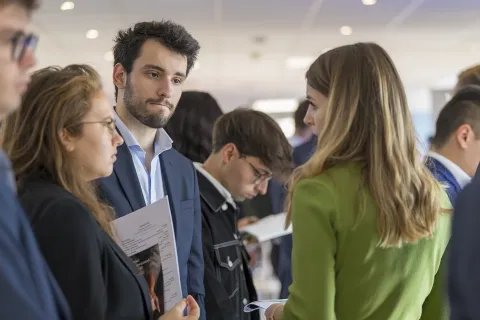 Group of people in business attire talking and exchanging documents during a professional networking event indoors.