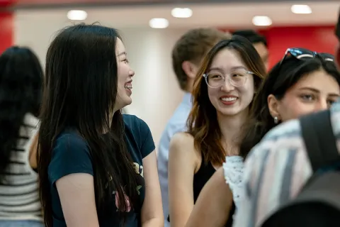 People standing and talking together during an indoor student networking event, with others visible in the background.
