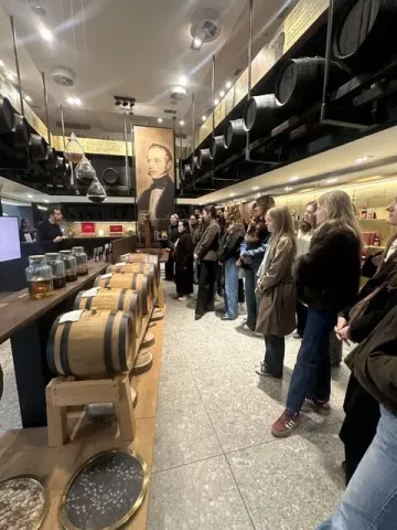  Group of people standing in an indoor visit or training space, listening to a presentation in front of aligned wooden barrels, within a professional environment focused on knowledge sharing.