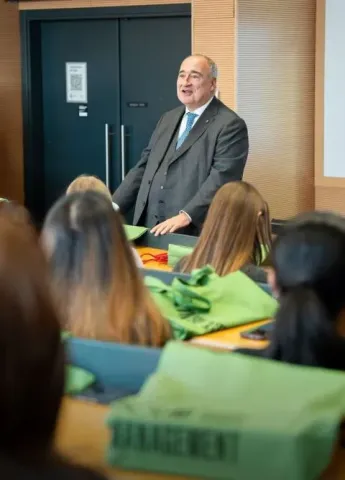  A speaker standing in front of an audience seated in a modern lecture room, with desks and presentation materials visible, in a higher education and academic training context.