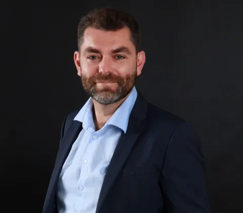 Portrait photographique d’une personne cadrée en buste, portant une veste sombre sur une chemise claire, avec une barbe, sur un fond uni sombre